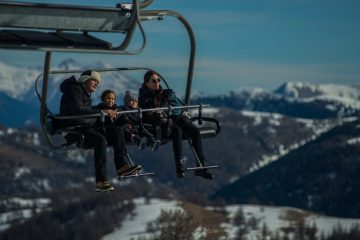 Family having fun on a ski lift with snow-covered mountains in the background during a sunny winter day.